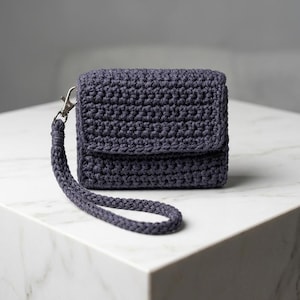 May include: A dark charcoal gray crochet wristlet pouch featuring a textured stitch pattern, a fold-over flap closure, and a matching braided wrist strap attached with a silver-toned metal lobster clasp. The accessory has a compact rectangular shape.