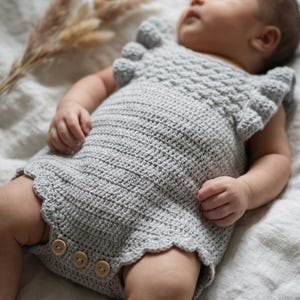 May include: A light gray crocheted baby romper featuring a textured bodice, ruffled sleeves, and a scalloped leg opening. The garment is secured at the bottom with three round wooden buttons, showcasing a detailed stitch pattern throughout the piece.