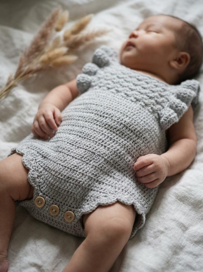 May include: A light gray crocheted baby romper featuring a textured bodice, ruffled sleeves, and a scalloped leg opening. The garment is secured at the bottom with three round wooden buttons, showcasing a detailed stitch pattern throughout the piece.
