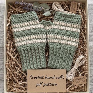 May include: A pair of sage green and cream striped crochet fingerless gloves with ribbed cuffs, presented in a gift box with brown paper filler, a card reading "Crochet hand cuffs pdf pattern," and tags labeled "HANDMADE WITH LOVE."