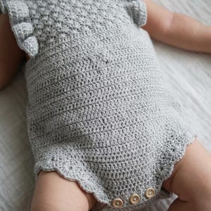 May include: A light gray crochet romper featuring a textured shell stitch pattern on the bodice, ruffled sleeves, and a scalloped leg opening. The garment includes three small, round, light-colored wooden buttons at the bottom crotch area.