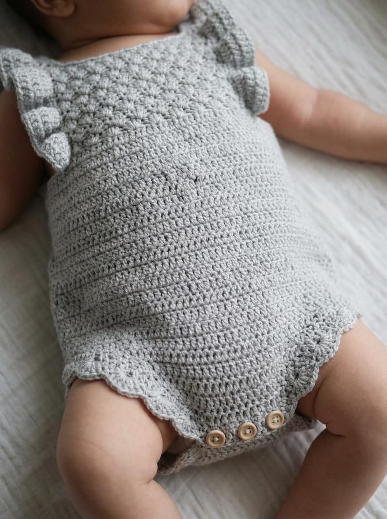 May include: A light gray crochet romper featuring a textured shell stitch pattern on the bodice, ruffled sleeves, and a scalloped leg opening. The garment includes three small, round, light-colored wooden buttons at the bottom crotch area.