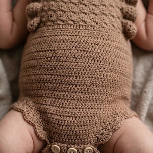 May include: A light brown crochet romper featuring a textured bodice with ruffled shoulder straps and a scalloped leg opening. The garment is secured at the bottom with three light-colored circular buttons, showcasing a detailed stitch pattern.
