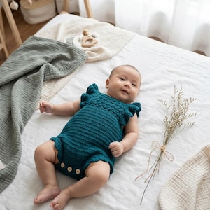 May include: A dark teal crochet baby romper featuring ruffled sleeves, a textured bodice, and three light-colored wooden buttons at the bottom. The garment is sleeveless with a round neckline and a ribbed knit pattern throughout the main body.