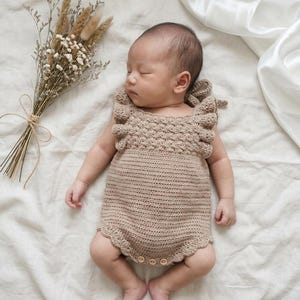 May include: A beige crochet baby romper featuring a textured bodice with ruffled shoulder straps, a smooth knit lower section, and a scalloped leg opening. The garment includes three small wooden buttons at the bottom for closure and ties at the back.