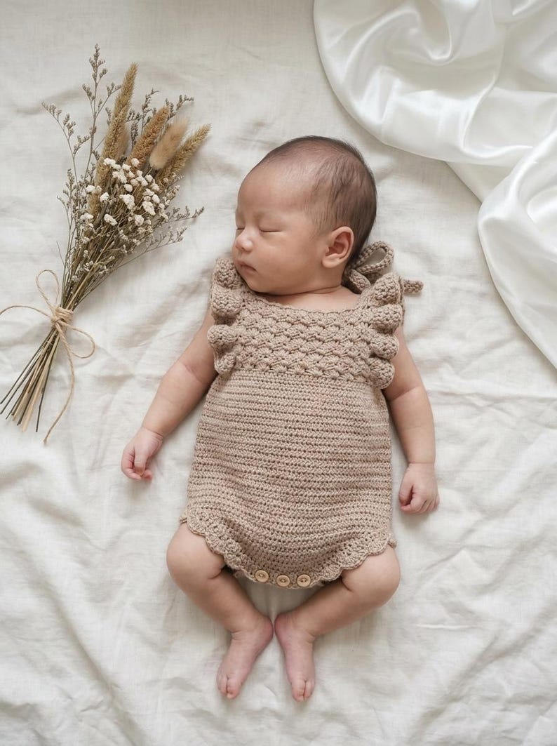 May include: A beige crochet baby romper featuring a textured bodice with ruffled shoulder straps, a smooth knit lower section, and a scalloped leg opening. The garment includes three small wooden buttons at the bottom for closure and ties at the back.