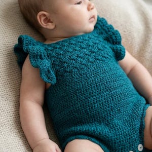 May include: A teal crochet romper featuring ruffled sleeves and a textured stitch pattern. The garment includes three white buttons at the bottom closure, providing a secure fit. The piece is crafted with a deep, solid teal yarn throughout.