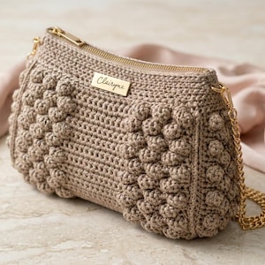 Può includere: Una borsa a mano all'uncinetto beige con cerniera e tracolla a catena dorate. La borsa presenta un design a punto nocciolina strutturato e una targhetta dorata con la scritta "Clairyne". La borsa è esposta su una superficie di marmo.