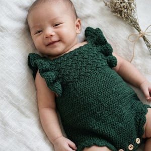 May include: A dark forest green crocheted baby romper featuring ruffled sleeves with a textured bobble stitch pattern and three light-colored wooden buttons at the bottom leg opening for closure. The garment has a scalloped edge along the leg holes.