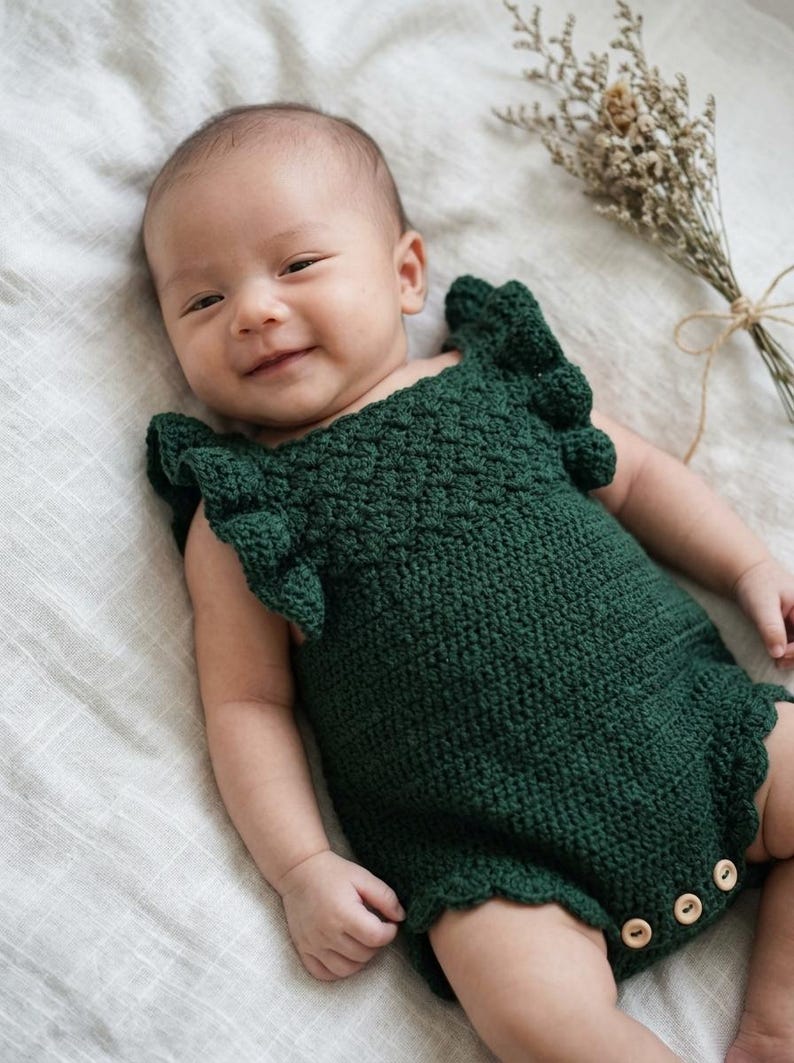 May include: A dark forest green crocheted baby romper featuring ruffled sleeves with a textured bobble stitch pattern and three light-colored wooden buttons at the bottom leg opening for closure. The garment has a scalloped edge along the leg holes.