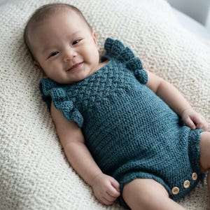 May include: A dark teal crochet romper featuring a textured diamond pattern on the bodice, ruffled sleeves, and three small wooden buttons at the bottom leg opening. The garment is crafted with a visible knit stitch and has a fitted silhouette.