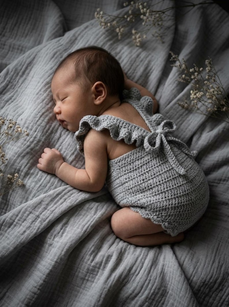 May include: A light gray crocheted romper featuring ruffled shoulder straps and a tie-back closure. The garment is crafted with a textured stitch pattern and scalloped leg openings, providing a soft and flexible fit for a newborn infant.