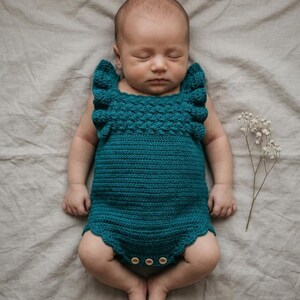 May include: A teal crochet baby romper featuring a textured bodice with ruffled shoulder straps and a scalloped leg opening. The garment is secured at the bottom with three small, light-colored wooden buttons, providing a functional closure.