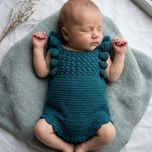 May include: A teal crochet baby romper featuring a textured bobble stitch bodice, ruffled shoulder straps, and a scalloped leg opening. The garment is crafted with a solid stitch pattern on the lower section and includes small circular buttons.