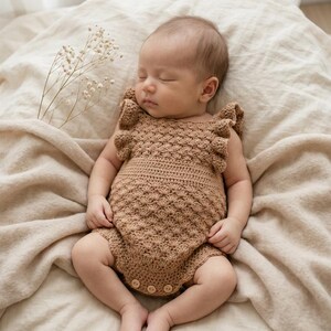 May include: A tan crochet romper featuring a textured shell stitch pattern, ruffled shoulder straps, and three small wooden buttons at the bottom. The garment is crafted with a fitted waistband and a scalloped edge design, providing a detailed look.