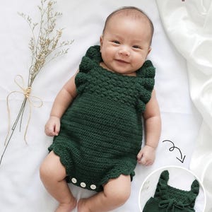 May include: A dark green crocheted baby romper featuring ruffled shoulder straps, a textured bodice, and a scalloped leg opening. The garment includes three small white buttons at the bottom. The text "Baby Romper" is visible in the upper right corner.