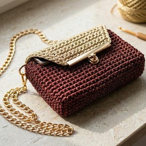 Puede incluir: Un bolso de mano de ganchillo burdeos con una cadena dorada y un cierre dorado. La solapa es de color oro claro. Una cadena dorada está sujeta al bolso. Una madeja de hilo dorado y un ganchillo están en el fondo.