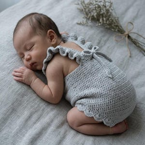 May include: A light gray crocheted baby romper featuring ruffled shoulder straps, a scalloped hemline, and a tie closure at the back. The garment is crafted with a textured stitch pattern and is designed to fit an infant, covering the torso and hips.