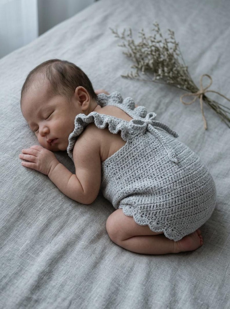 May include: A light gray crocheted baby romper featuring ruffled shoulder straps, a scalloped hemline, and a tie closure at the back. The garment is crafted with a textured stitch pattern and is designed to fit an infant, covering the torso and hips.