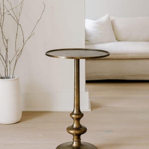May include: A round side table with a metallic antique brass finish. The table features a flat circular top supported by a slender, decorative pedestal base that flares out into a wide, stable circular foot at the bottom.