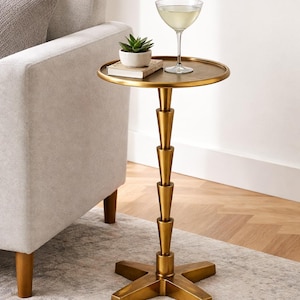 May include: A round gold metal side table with a slender, stacked conical pedestal base and a four-point cross base. The tabletop features a smooth surface holding a small potted green succulent plant and a clear glass cocktail coupe.