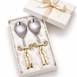 Könnte beinhalten: Ein Set aus zwei Servierlöffeln mit polierten silbernen Schalen und goldfarbenen Schleifen-Griffen, präsentiert in einer weißen Geschenkbox mit Satinband. Die Löffel liegen in einem weißen Satinfutter.