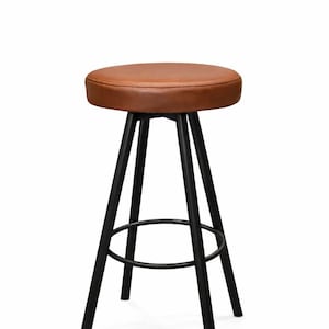 Puede incluir: Taburete de bar con asiento de cuero marrón y estructura de metal negro. El asiento redondo mide aproximadamente 30 cm de diámetro. El taburete tiene un reposapiés y tres patas que se inclinan hacia afuera. Fondo blanco.