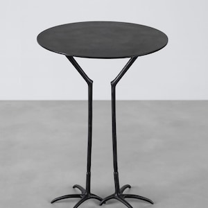 May include: A black metal side table with a round top. The table has a unique design, with two legs that split into a bird-like foot base. The table's height is approximately 71 cm.