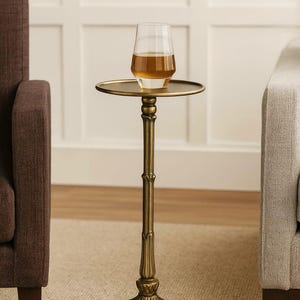 May include: A small, antique gold side table with a round top and a decorative stem. A glass of amber liquid sits on the table. The table is next to a brown upholstered armchair.
