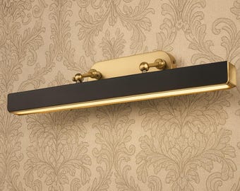 May include: A rectangular wall-mounted light fixture with a black center and gold-colored trim and accents. The light has a warm glow and is mounted on a gold-colored base. The fixture has two decorative knobs.