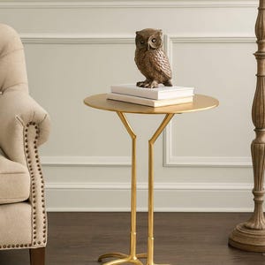 Può includere: Un tavolino dorato con piano rotondo e base a tre gambe. Una statuetta di gufo è appoggiata su due libri bianchi sul tavolo. Il tavolo è accanto a una poltrona beige e una lampada da terra in legno.
