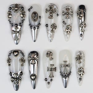 May include: A set of ten long, pointed artificial nails. Each nail is adorned with silver-colored cross and floral embellishments. Some nails feature small, round studs. The nails have a clear or silver base.