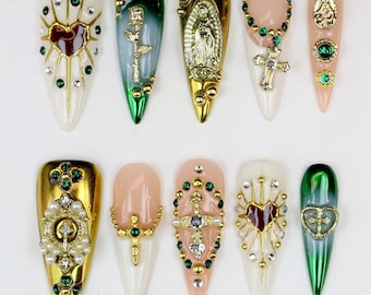 May include: A collection of ten artificial nails with various designs. The nails feature a range of colors, including gold, green, white, and pink. Embellishments include crosses, hearts, and religious figures, with rhinestones and pearls.
