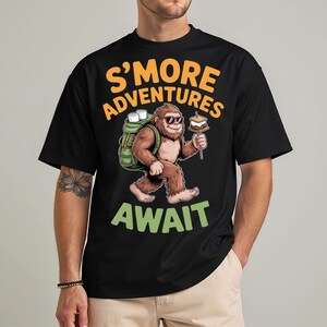 Puede incluir: Camiseta negra con un gráfico de un personaje de Bigfoot con gafas de sol y una mochila, sosteniendo un s'more. El texto dice "S'MORE ADVENTURES AWAIT" en naranja y verde.