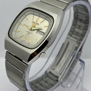 Puede incluir: Un reloj de pulsera automático Seiko plateado con esfera rectangular. La esfera es plateada clara con agujas e indicadores dorados. El día y la fecha se muestran a las 3. La correa es de acero inoxidable.