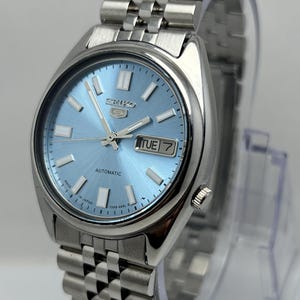 May include: A silver-tone stainless steel Seiko 5 automatic wristwatch featuring a light blue sunray dial with silver baton indices, luminous hands, and a day-date window at the three o'clock position, paired with a multi-link metal bracelet.