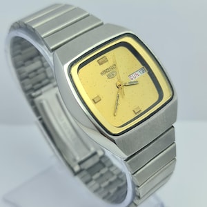 May include: A vintage Seiko 5 wristwatch with a gold-coloured square dial and a silver-tone stainless steel band. The watch face displays the day and date. The watch is displayed on a clear stand.