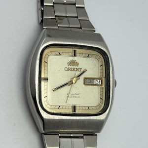 May include: Silver-toned Orient watch with a rectangular face and a matching metal band. The watch face is cream-colored with gold-toned hands and markers. The day and date are displayed in a small window. The watch is a vintage style.