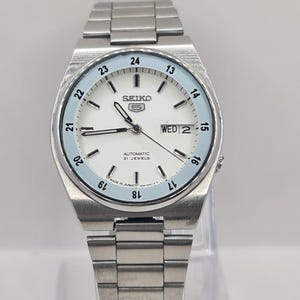 May include: A silver-toned Seiko 5 automatic wristwatch with a white face and a light blue outer ring. The watch displays the day and date, with the text "WED 2" visible. The watch band is made of silver metal links.