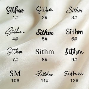 K&ouml;nnte beinhalten: Zw&ouml;lf schwarze Textdesigns auf wei&szlig;em Hintergrund. Das Wort "Sithm" ist in einer Schreibschrift geschrieben, jeweils mit einer Nummer von 1 bis 12. Die Buchstaben "SM" sind ebenfalls enthalten. Der Hintergrund ist ein weicher, strukturierter Stoff.