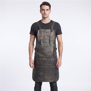 Könnte beinhalten: Eine dunkelbraune Lederschürze mit verstellbaren Schultergurten und Metallschnallen, zwei Brusttaschen, zwei seitlichen Metallringen und einer großen unteren Tasche mit mehreren Werkzeugfächern. Die Oberfläche hat ein Used-Finish.