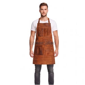 May include: A brown leather apron featuring multiple utility pockets and tool loops on the front. The apron has adjustable shoulder straps with metal buckles and is worn over a white t-shirt and dark grey jeans. The text Leather Gifter is visible.