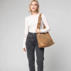 May include: A tan suede hobo bag with a wide shoulder strap and a rectangular buckle detail. The bag features a soft, slouchy construction with visible seam stitching. The text Leather Gifter is overlaid on the center of the tan suede bag.