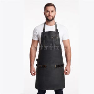 May include: A black leather apron featuring adjustable shoulder straps with metal buckles, two upper chest pockets, and a horizontal row of small loops across the waist area. The text Leather Gifter is printed in white across the centre chest.