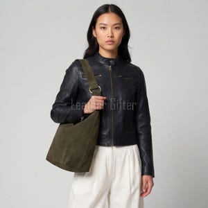 Può includere: Una borsa hobo in camoscio verde oliva scuro con un'ampia tracolla e un anello metallico circolare. La borsa è indossata a spalla insieme a una giacca in pelle nera con chiusura a zip frontale e due tasche orizzontali sul petto.