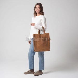 Op de afbeelding: Een lichtbruine leren tas met een grote vierkante voorzak en twee schouderbanden. De tas heeft een gladde, matte afwerking en een rechthoekig silhouet. De tekst "Leather Gifter" is zichtbaar in een lichtgrijs lettertype op de tas.
