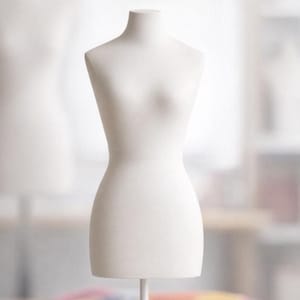 Può includere: Un manichino da sartoria bianco, dalla forma femminile e slanciata. Presenta collo, spalle, busto, vita e fianchi, sostenuto da un sottile supporto in metallo bianco. Lo sfondo è sfocato, suggerendo un ambiente da studio. Ideale per esporre abiti.
