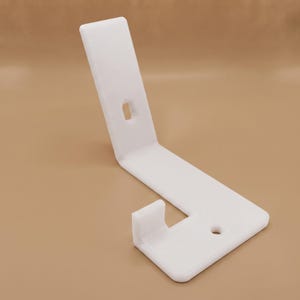 May include: A white L-shaped plastic bracket featuring a flat base with a circular mounting hole, an upward-angled arm with a rectangular slot, and a small perpendicular hook protruding from the base for securing items in place.