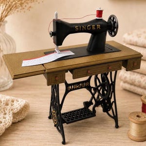 Op de afbeelding: Een miniatuur Singer naaimachine met een zwart metalen frame met gouden letters, een goudkleurig tafeloppervlak met kleine lades en een pedaalonderstel. Een rode garenklos staat bovenop en een stuk witte stof ligt onder de naald.