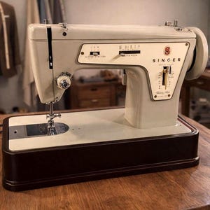 Könnte beinhalten: Vintage Singer Nähmaschine in Creme- und Brauntönen. Die Maschine hat das Singer-Logo und verschiedene Knöpfe und Einstellungen. Der Sockel ist dunkelbraun und steht auf einer Holzoberfläche.